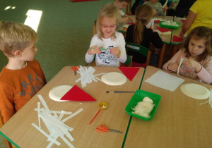 Dzieci przy stolikach wykonują portret Mikołaja z papierowych talerzyków doklejając różne elementy.